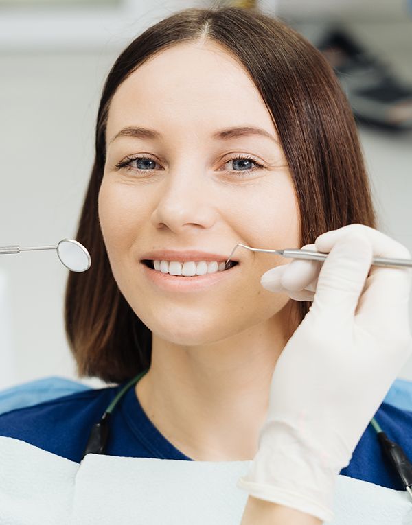 Chica morena sonriendo en la silla del dentista. Se ven las manos del dentista sosteniendo instrumental para la revisión dental.