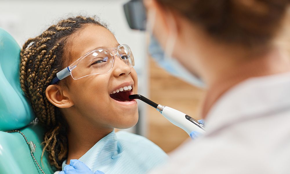 Niña pequeña con gafas protectoras abriendo la boca mientras el dentista le realiza el tratamiento.